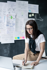 Young contemporary female office worker sitting on desk and making notes in copybook while planning work
