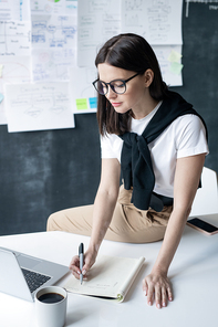 Young contemporary female office worker sitting on desk in front of laptop and making notes in copybook