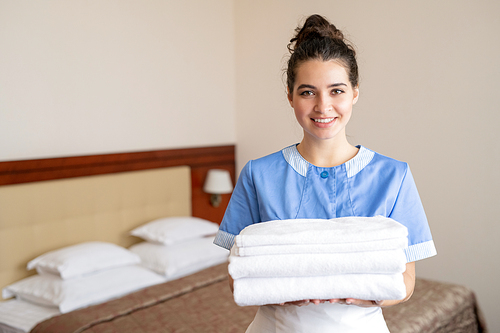 Pretty young smiling chamber maid in uniform holding stack of white clean soft towels while standing in front of camera
