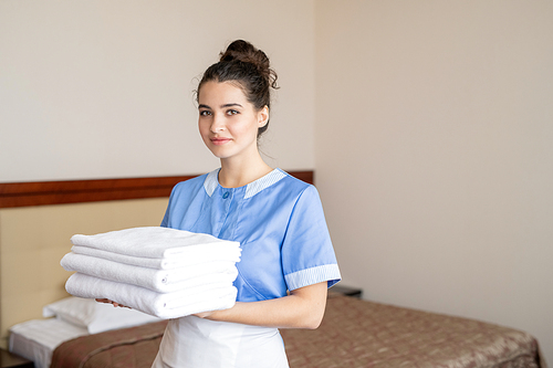 Pretty young woman in uniform of chamber maid looking at you while carrying stack of white fresh soft towels