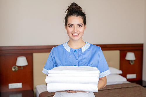 Pretty young chamber maid holding stack of white clean towels while doing her work in hotel room