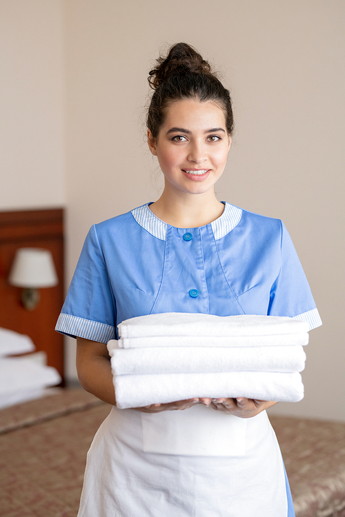 Pretty girl in blue uniform and white apron looking at you while holding stack of fresh white towels during work in hotel