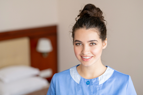 Happy young brunette female in blue uniform of chamber maid standing in hotel room and looking at you