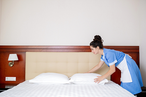 Pretty young woman in uniform of room maid standing by bed and changing pillows in the morning