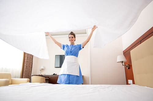 Pretty young chamber maid in uniform stretching white clean linen over bed while working in hotel room