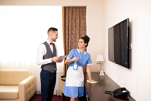 Young chamber maid with detergent and duster and porter with touchpad interacting in one of hotel rooms at work