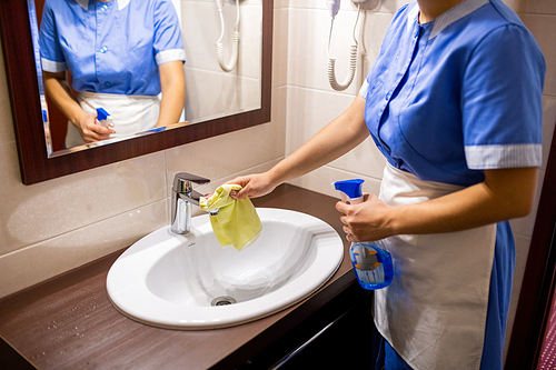Young chamber maid in uniform standing in front of mirror in bathroom and cleaning sink with detergent
