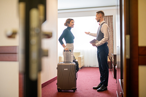 Pretty young elegant woman with luggage standing in hotel room and talking to porter welcoming her