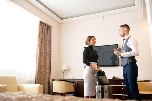 Young elegant porter with touchpad talking to pretty businesswoman with baggage while both standing in hotel room