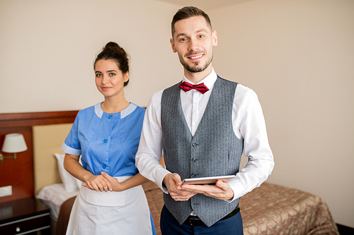 Young successful porter and pretty chamber maid in uniform standing in front of camera in hotel room