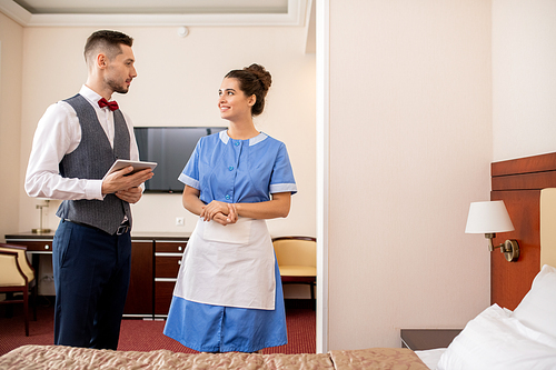 Cheerful young room maid in uniform talking to elegant porter with touchpad while working in hotel room
