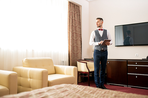 Contemporary young elegant porter standing in one of hotel rooms, looking at white leather armchair and entering information in touchpad