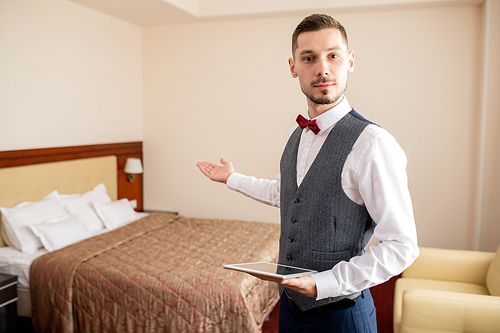Young professional in elegant clothes holding touchpad and pointing at bed while standing in front of camera in hotel room