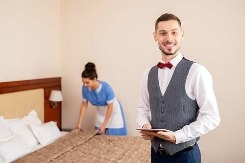 Young successful hotel staff with toothy smile standing in front of camera on background of chamber maid making bed