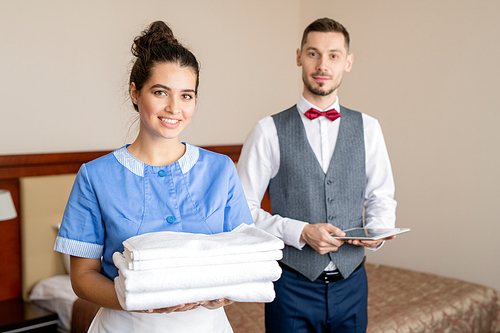 Pretty young smiling chamber maid with stack of clean white towels standing in front of camera with porter behind