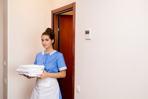 Young brunette chamber maid in uniform carrying stack of white fresh towels while going out of one of hotel rooms