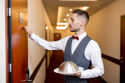Young elegant waiter knocking on wooden door of hotel room while holding food on cloche for one of guests