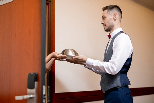 Young hospitable waiter in uniform passing cloche with breakfast or lunch to female guest by open door of hotel room