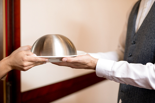 Hands of young woman taking cloche with restaurant food from elegant waiter through open door of hotel room