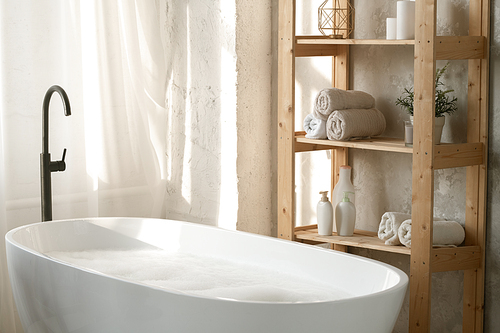 Large porcelain white bathtub filled with water and foam by wooden shelves with rolled towels and plastic jars against wall in bathroom