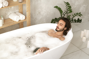 Young relaxed man with closed eyes lying in white bathtub filled with hot water and foam with wooden shelves near by