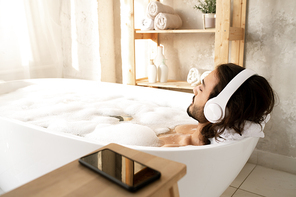 Young man with headphones lying in white bathtub filled with hot water and foam and enjoying relaxing music