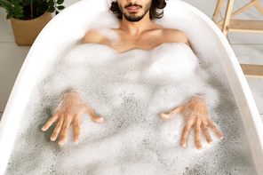 Young man lying in white bathtub filled with hot water and touching foam by his hands while relaxing