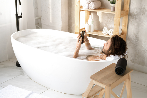Young restful man using smartphone while lying in bathtub filled with water and foam in the corner of bathroom