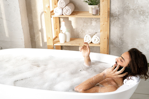 Young restful man talking by smartphone while lying in bathtub filled with water and foam in bathroom