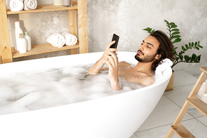Young naked man watching video or scrolling in smartphone while lying in bathtub filled with water and foam in bathroom