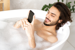 Cheerful young man with smartphone watching video or making selfie while lying in bathtub filled with water and foam