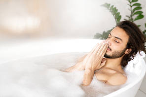 Young serene man meditating while enjoying hot bath with foam with his hands put together by face