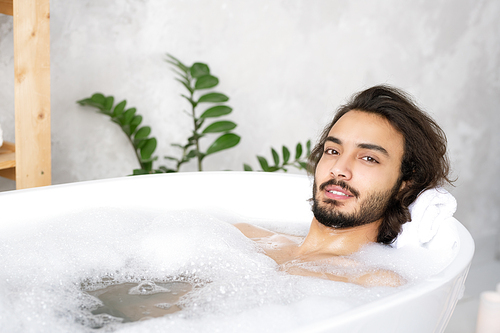 Young serene bearded man relaxing while lying in bath with hot water and foam in front of camera