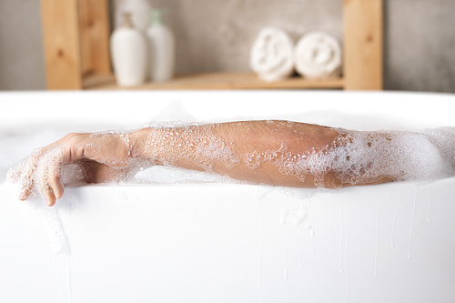 Arm of young man relaxing in white porcelain bath with foam on background of shelf with towels and body care products