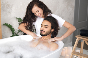 Pretty young woman making massage of neck and shoulders to her husband relaxing in bath with hot water and foam