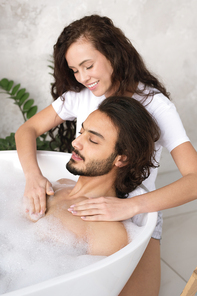 Pretty young smiling woman making massage of chest to her husband relaxing in bath with hot water and foam