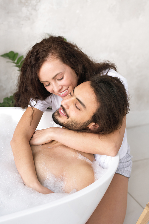 Happy young affectionate woman embracing her relaxed husband enjoying bath with hot water and foam