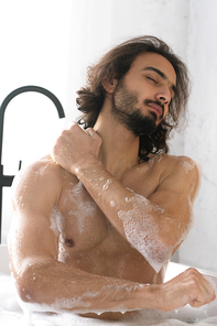 Young muscular man with closed eyes washing his body while enjoying bath with hot water and foam