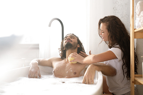 Careful young woman washing body of her relaxed husband with sponge while sitting by bathtub