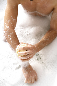 Overview of shirtless young man with sponge washing his arms while sitting in bath with hot water and foam
