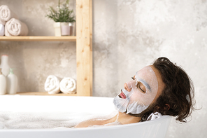 Serene and relaxed young man with face mask enjoying hot bath with foam while spending time in bathroom