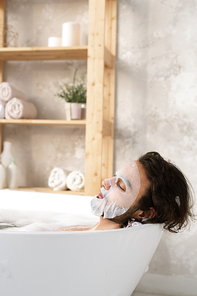 Relaxed young man having face mask while lying in bath with hot water and foam on background of shelf with self-love items