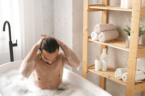 Wet young muscular man washing his hair while having bath with foam by shelves with self-love items