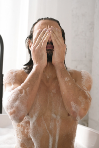 Wet young muscular man keeping his hands by face while washing his body in bath with hot water and foam