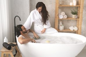 Happy young woman in white bathrobe sitting on bathtub and talking to her husband enjoying bath with foam