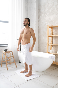 Young shirtless man with soft white towel on hips standing on small rug after having hot bath with foam