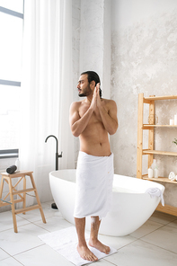Young shirtless muscular man with soft white towel on hips standing on the floor of bathroom after having hot bath with foam