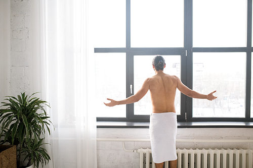 Rear view of young man with white towel on hips outstretching his arms while looking through window after having bath