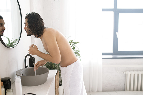 Shirtless young man with white towel on hips looking in mirror while washing his face over sink in bathroom