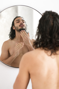 Shirtless young man looking at his beard in mirror while going to shave after having bath in the morning